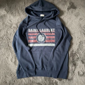 Mörkblå Saint Laurent hoodie - Snygg mörkblå hoodie från Saint Laurent med tryck i rött och vitt framtill. Tröjan har huva med snörning och en klassisk känguruficka. Perfekt för dig som gillar streetwear och vill ha något med coolt print.