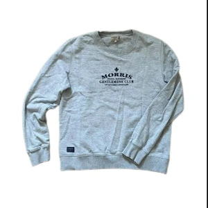 Grå sweatshirt från Morris, M - Snygg grå sweatshirt från Morris med mörkblå tryck på bröstet där det står 'Gentlemens Club'. Tröjan har rund hals, ribbade muddar och är långärmad. Perfekt för dig som gillar klassisk och stilren design med en sportig touch. Sparsamt använd och inga defekter 