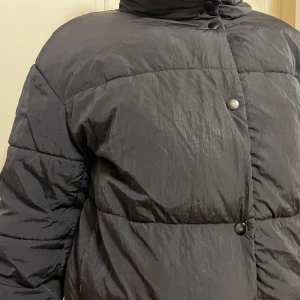 Svart pufferjacka från Zara - Svart pufferjacka från Zara med hög krage och tryckknappar framtill. Jackan har en ingömd hoodie! Jackan har en loose passform och är tillverkad i syntetmaterial som ger en glansig finish. Perfekt för kalla dagar med sin vadderade design och långa ärmar.