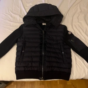 Mörkblå  Moncler pufferjacka med huva - Svart pufferjacka från Moncler i storlek M med stickade ärmar och detaljer. Jackan har huva, ribbade muddar, dragkedja framtill och Moncler-logga på ärmen. Snygg kombination av quiltad polyester och stickad material, samt praktisk ficka med dragkedja på ärmen.känns som en S