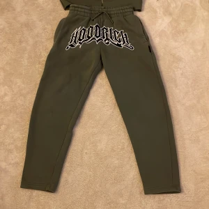 Hoodrich hoodie i olivgrönt med tryck - Olivgrön hoodie från Hoodrich med stor vit och svart logotyp på bröstet och huva med tryck. Tröjan har dragkedja, känguruficka och ribbade muddar. Materialet är mjuk bomull och polyester, perfekt för chill eller streetwear. Snyggt matchad med tillhörande byxor.