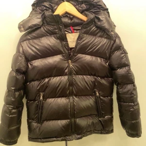 Moncler Jacka - Svart pufferjacka från Moncler med glansig finish, huva och dragkedjor fram samt på fickorna. Jackan har Moncler-logga på ärmen och quiltad design som ger en schysst streetstil. Perfekt för kalla dagar när du vill hålla värmen och se fresh ut. Storlek 160 cm, priset är inte hugget, tveka inte på att skriva för fler frågor och funderingar🙌