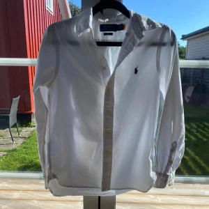 Vit skjorta från Polo Ralph Lauren - Klassisk vit skjorta från Polo Ralph Lauren i slim fit-modell. Skjortan har långa ärmar, knappar framtill och den ikoniska broderade loggan på bröstet. Tillverkad i bomull för en fräsch och stilren look. Är i fint skick och är köpt på NK.