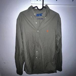 Olivgrön skjorta från Ralph Lauren - Olivgrön skjorta från Ralph Lauren med klassisk orange broderad logga på bröstet. Skjortan har lång ärm, knappar framtill och button-down krage. Tillverkad i bomull för en skön och avslappnad känsla.