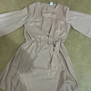 Rosa glittrig kortklänning med bälte - Superfin rosa kortklänning med glittriga vertikala ränder och långärmade ärmar. Klänningen har ett matchande bälte i midjan och en lätt, flowy känsla. Perfekt för dig som vill sticka ut med en elegant men chill look.