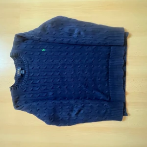 Marinblå kabelstickad tröja Polo Ralph Lauren - Kabelstickad marinblå tröja från Polo Ralph Lauren i storlek M. Tröjan har rund halsringning, lång ärm och klassisk grön broderad logga på bröstet. Tillverkad i mjuk bomull och har ribbade muddar vid ärmslut och nederkant. Strl L i barn strl vilket motsvarar M i herr strl.