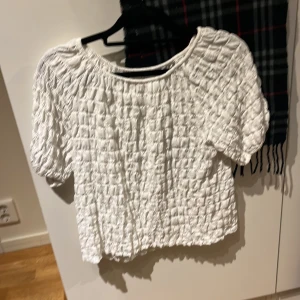 Vit strukturerad blus från Zara - Säljer en vit blus från Zara med korta puffärmar och rund hals. Toppen har ett coolt rutigt, strukturerat tyg som ger en unik look. Perfekt för dig som gillar trendiga och bekväma plagg med lite extra detalj.