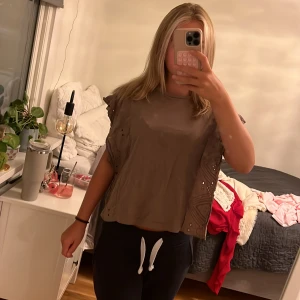 Brun blus med volang från Kappahl - Säljer en brun blus från Kappahl i storlek S. Toppen har korta ärmar med volang och snygga broderade detaljer längs sidorna. Den är loose fit och tillverkad i mjuk bomull, perfekt för en chill och trendig look.