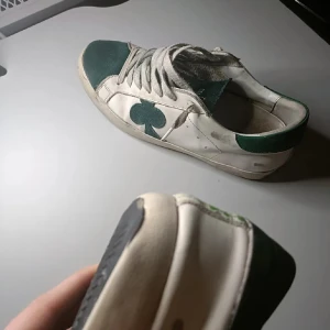 Gröna och vita sneakers The Gallerist - Sneakers i vitt och grönt från The Gallerist med snygga klöverdetaljer på sidan och hälen. Ovandel i skinn och mocka, gröna partier på tå, plös och häl. Klassisk snörning och platt sula. Perfekta för dig som gillar unika detaljer.