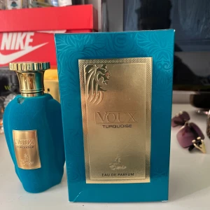 Voux Turquoise Eau de Parfum - Parfymen Voux Turquoise Eau de Parfum kommer i en lyxig turkos flaska med sammetsliknande yta och guldfärgad metallkork. Flaskan har en elegant, rundad form och en guldig etikett med lejonmotiv. Förpackningen matchar flaskans färg och har detaljer i guld.