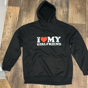Svart hoodie med I ❤️ MY GIRLFRIEND-print - Svart hoodie med stor vit text 'I ❤️ MY GIRLFRIEND' och rött hjärta på bröstet. Tröjan har huva med snörning och en klassisk magficka. Materialet är mjukt och känns som polyester, perfekt för chill dagar. Långärmad och loose fit.