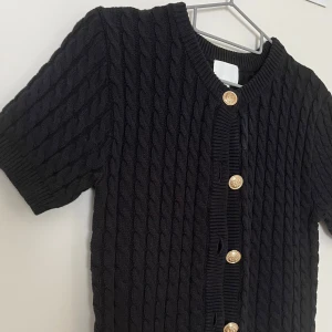 Svart kabelstickad kofta med guldknappar - Svart kabelstickad kofta med korta ärmar och snygga guldknappar framtill. Tröjan har rund hals och ribbade muddar vid ärmslut och nederkant. Perfekt för dig som gillar klassisk stil med en modern twist.