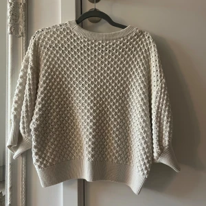 Vit stickad tröja med strukturmönster - Säljer en vita stickad tröja med snyggt strukturmönster och breda ribbade muddar vid ärmslut och nederkant. Tröjan har en loose passform och rund halsringning. 