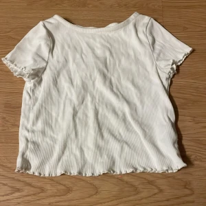 Vit ribbad topp från H&M - Vit ribbad topp från H&M med korta ärmar och söt volangkant vid ärmslut och nederkant. T-shirten har en enkel, rak passform och är tillverkad i mjuk bomull. Perfekt för en clean och fräsch look.