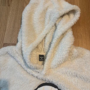 Fluffig hoodie med nalle från Manfinity - Supermjuk och fluffig vit hoodie från Manfinity med stor huva och nallebjörn med hörlurar tryckt på bröstet och ärmen. Perfekt för dig som vill ha en mysig och chill vibe. Ribbstickade muddar och magficka framtill.