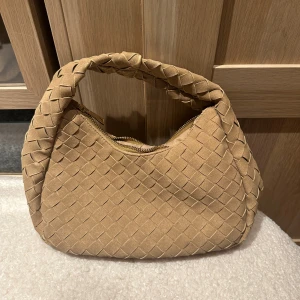 Beige flätad axelväska i mocka - Snygg beige axelväska med flätad design och mjukt material från Glitter. Väskan har ett kort, vadderat handtag och stängs med dragkedja. Perfekt för dig som gillar trendiga accessoarer med unik struktur.