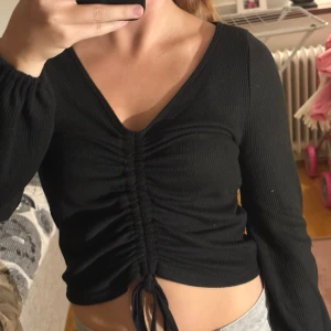 Svart croppad ribbad topp med snörning - Säljer en svart croppad topp med lång ärm och snygg v-ringning. Toppen har ribbad struktur och snörning framtill som ger en cool rynkad effekt. Perfekt att styla med jeans eller kjol för en trendig look. Materialet känns mjukt och stretchigt.