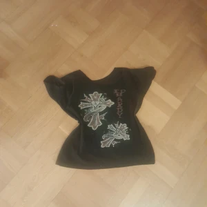 Svart Ed Hardy topp med kors och nitar - Cool svart topp från Ed Hardy med stora korsmotiv framtill, dekorerade med glittriga nitar och färgade detaljer. Toppen har korta ärmar och figurnära passform. Snyggt tryck med Ed Hardy-logga både fram och bak. Perfekt för dig som gillar statement-plagg. PRIS KAN DISKUTERAS