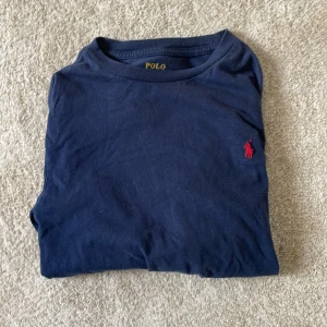 Mörkblå långärmad t-shirt Polo Ralph Lauren - Snygg mörkblå långärmad t-shirt från Polo Ralph Lauren med klassisk röd broderad logga på bröstet. Tillverkad i mjuk bomull med rund hals och normal passform. Perfekt för dig som gillar stilrena och tidlösa plagg.