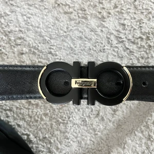 Svart Ferragamo bälte 90cm - Snyggt svart skärp från Ferragamo i skinn med ikoniskt dubbelringat spänne i svart och guld. Bältet har en stilren och lyxig känsla, perfekt för att lyfta vilken outfit som helst. Klassisk design med Ferragamo-logga på spännet storlek är mellan 90 cm till 100cm.