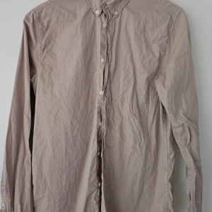 Beige skjorta från Knowledge Cotton - Stilren beige skjorta från Knowledge Cotton Apparel i storlek S. Skjortan har klassisk krage, långa ärmar och knäppning framtill med vita knappar. Tillverkad i mjuk bomull och passar perfekt till en avslappnad men snygg look. I bra skick använd enstaka gång, säljes då jag lagt på mig och den inte passar längre. Hel och ren, lär behöva en strykning.  (Katt finns i hemmet)