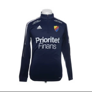Marinblå Djurgården fotbollströja Adidas M - Snygg marinblå Djurgården fotbollströja från Adidas med lång ärm och halv dragkedja. Tröjan har klubbmärke på bröstet, vita tryck med sponsorer och klassiska Adidas-ränder på axlarna. Tillverkad i 100% återvunnen polyester (Primegreen). Perfekt för träning eller match.