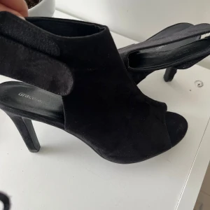 Svarta mocka klackskor från Graceland - Svarta klackskor i mocka från Graceland med öppen tå och hög klack. Snygg omlott-design med kardborreknäppning runt ankeln. Perfekta för dig som vill ha en stilren och trendig look med lite extra edge.