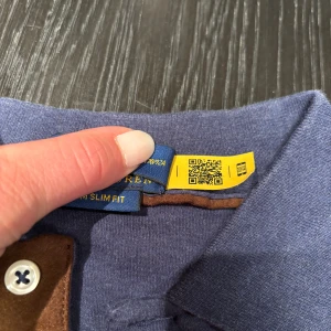 Blå långärmad pikétröja Polo Ralph Lauren - Snygg blå långärmad pikétröja från Polo Ralph Lauren i custom slim fit. Tröjan har klassisk krage, bruna detaljer vid knäppningen och tre knappar framtill. Ikonisk broderad logga på bröstet. Perfekt för dig som gillar stilren och sportig look.