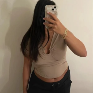 Beige croppad topp med v-ringning - Säljer en beige croppad topp med djup v-ringning och snygg rynkdetalj framtill. Toppen är tight och har korta ärmar, perfekt för en trendig look. Materialet känns mjukt och stretchigt, vilket gör den bekväm att bära.