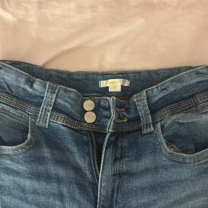 Blå jeans från Gina Tricot med dubbla knappar - Säljer ett par klassiska blå jeans från Gina Tricot med dubbla silverfärgade knappar och dragkedja. Jeansen har fem fickor, bälteshällor och snygga sömdetaljer på bakfickorna. Perfekta till din vardagsstil! Använt en gång!