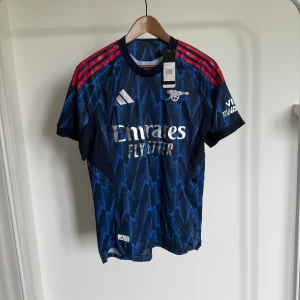 Arsenal 25/26 Adidas blå bortatröja M - Snygg Arsenal bortatröja från Adidas i mörkblått med elektriskt blått blixtmönster och röda ränder på axlarna. Tröjan har korta ärmar, rund hals och är tillverkad i lätt, ventilerande material. Stora loggor från Adidas och Emirates på bröstet.