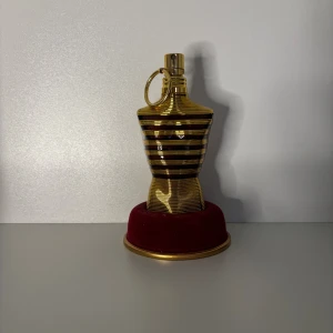 Jean Paul Gaultier Le Male Parfym sampel  - Unik parfymflaska i form av en manlig torso i guld med svarta ränder och en stor ringdetalj vid halsen. Flaskan står på en rund, vinröd sammetsliknande bas med guldkant. Perfekt för dig som gillar statement-detaljer.