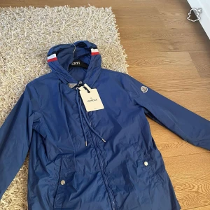 Blå vindjacka från Moncler med huva - Blå vindjacka från Moncler med huva och dragkedja framtill. Jackan har vita och röda detaljer på huvan, två fickor med knappar och Moncler-logga på ärmen. Tillverkad i lätt polyester, perfekt för blåsiga dagar. Snygg och sportig look.