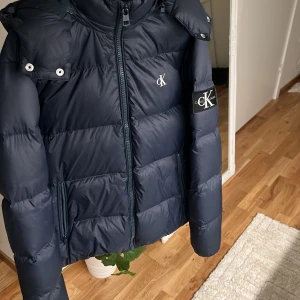Mörkblå pufferjacka från Calvin Klein - Snygg mörkblå pufferjacka från Calvin Klein Jeans med huva och dragkedja framtill. Jackan har logga på bröstet och ärmen, samt praktiska fickor med dragkedja. Perfekt för kalla dagar och riktigt stilren vibe.