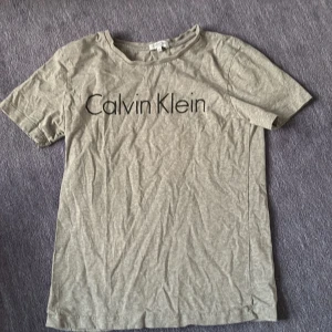 Grå Calvin Klein t-shirt med tryck - Snygg grå t-shirt från Calvin Klein med stort svart logotryck på bröstet. Klassisk rund halsringning och korta ärmar. Tillverkad i mjuk bomull som är skön mot huden. Perfekt för en clean och stilren look.