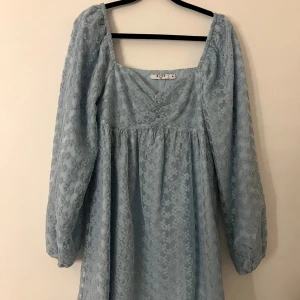 Ljusblå spetsklänning från NA-KD - Supersöt ljusblå kortklänning från NA-KD med långa puffärmar och v-ringning. Klänningen har ett blommigt spetsmönster och är lätt transparent med romantisk känsla.  Perflet för vår eller sommar, lite varmare pga de långa armarna. Räcker till lite kortare än mitten på låren. Använd en dag på ett bröllop.