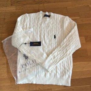 Vit stickad tröja  - Vit kabelstickad tröja  med klassisk rund halsringning och broderad logga på bröstet. Tröjan har långa ärmar och ribbade muddar, perfekt för en stilren och tidlös look.