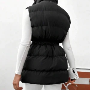 Svart lång dunväst med dragsko - Snygg svart dunväst med lång passform och markerad midja med dragsko. Västen har stora fickor med lock och coola metalldetaljer. Stängs med dragkedja framtill och är perfekt att slänga över en hoodie eller långärmad tröja.