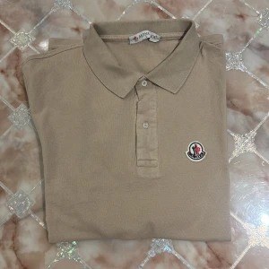 Beige Moncler pikétröja i bomull - Snygg beige pikétröja från Moncler i 100% bomull. Klassisk krage med knappar och broderad Moncler-logga på bröstet. Perfekt för en clean och stilren look, passar till jeans eller shorts. Skön passform och tidlös design.