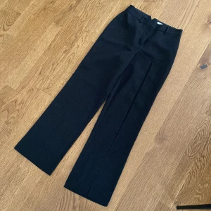 Svarta kostymbyxor från NA-KD ART - Stilrena svarta kostymbyxor från NA-KD ART i storlek 34. Byxorna har hög midja, vida ben och pressveck framtill. Perfekta för en clean och trendig look. Materialet känns mjukt och har snygg struktur. Bälteshällor upptill för extra stylingmöjligheter.