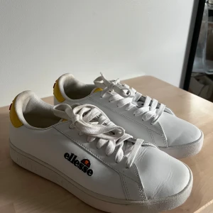 Vita sneakers från Ellesse med gula detaljer - Fräscha vita sneakers från Ellesse med snygga gula och orange detaljer på hälen och logga på sidan. Skorna har rund tå, platt sula och klassisk snörning. Använda endast en gång så iprincip i nyskick. 