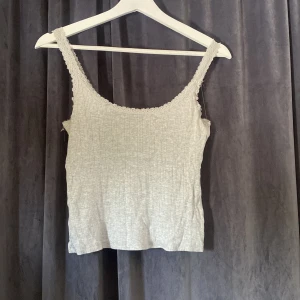 Ljusgrå ribbad linnetopp från 157 - Supersöt ljusgrå ribbad linnetopp från 157 med spetskant runt halsringningen och axelbanden. Toppen är ärmlös och har en mjuk, stretchig känsla tack vare bomullsmix med viskos. Perfekt att styla med jeans eller kjol för en chill look.