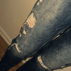 Blå ripped jeans med straight fit - Säljer ett par blå jeans med slitna detaljer och hål på knäna. Normal passform och rak siluett. Perfekta för dig som gillar en avslappnad och trendig look. 