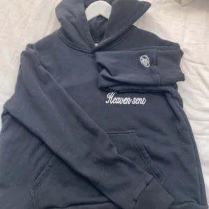 Svart hoodie Heaven Sent från Deema - Svart hoodie från Deema med broderad text 'Heaven sent' på bröstet och en liten logga på ärmen. Klassisk känguruficka framtill och huva. Perfekt för dig som gillar en clean och stilren look.