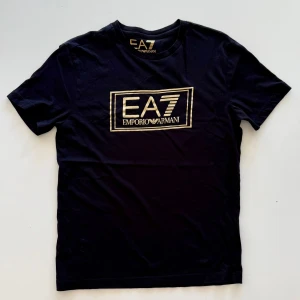 Svart EA7 Emporio Armani t-shirt - Svart t-shirt från EA7 Emporio Armani med guldig logga tryckt på bröstet. Klassisk passform med rund hals och korta ärmar. Perfekt för dig som gillar stilrena och exklusiva plagg med tydlig branding.