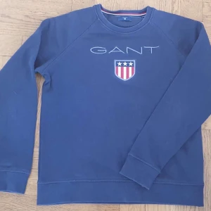 Blå sweatshirt från GANT - Säljer en klassisk blå sweatshirt från GANT med broderad logga och sköld på bröstet. Tröjan har rund halsringning och långa ärmar, perfekt för en chill och stilren look. Tillverkad i mjuk bomull för extra komfort.