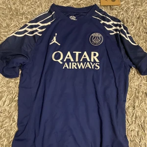 PSG x Jordan blå fotbollströja L - Blå PSG x Jordan fotbollströja i storlek L med vita detaljer och tryck. Tröjan har korta ärmar, Dri-Fit material och coola vita mönster på axlarna. Stort Qatar Airways-tryck på bröstet och PSG-logga. Perfekt för dig som vill sticka ut på planen.