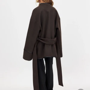 Brun omlottjacka från NA-KD - Snygg brun omlottjacka från NA-KD med breda ärmar och knytbälte i midjan. Jackan har svarta detaljer längs kanterna. Nypris är 1100:-. Använd någon gång - nyskick.