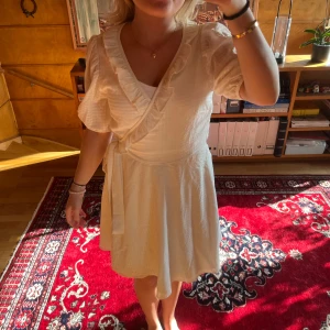 Vit omlottklänning med volang - Säljer en söt vit omlottklänning med puffiga korta ärmar och volangdetaljer längs v-ringningen. Klänningen har knyt i midjan och är tillverkad i ett lätt, mjukt tyg som känns luftigt. Perfekt för varma dagar och ger en romantisk vibe.