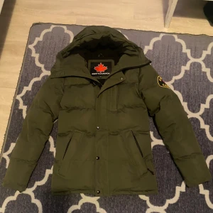 Grön pufferjacka från Zavetti Canada XS - Snygg mörkgrön pufferjacka från Zavetti Canada i storlek XS. Jackan har huva med fuskpäls, stora fickor och coolt märke på ärmen. Perfekt för kalla dagar med sin vadderade design och dragkedja framtill.